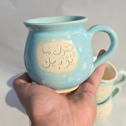 Hand holding a light blue ceramic mug with Arabic text on a plain background