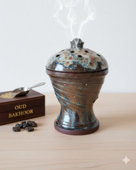 Ceramic incense burner with smoke, next to a box of oud bakhoor on a light surface.
