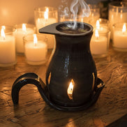 Black ceramic oil burner with smoke on a wooden surface surrounded by lit candles.