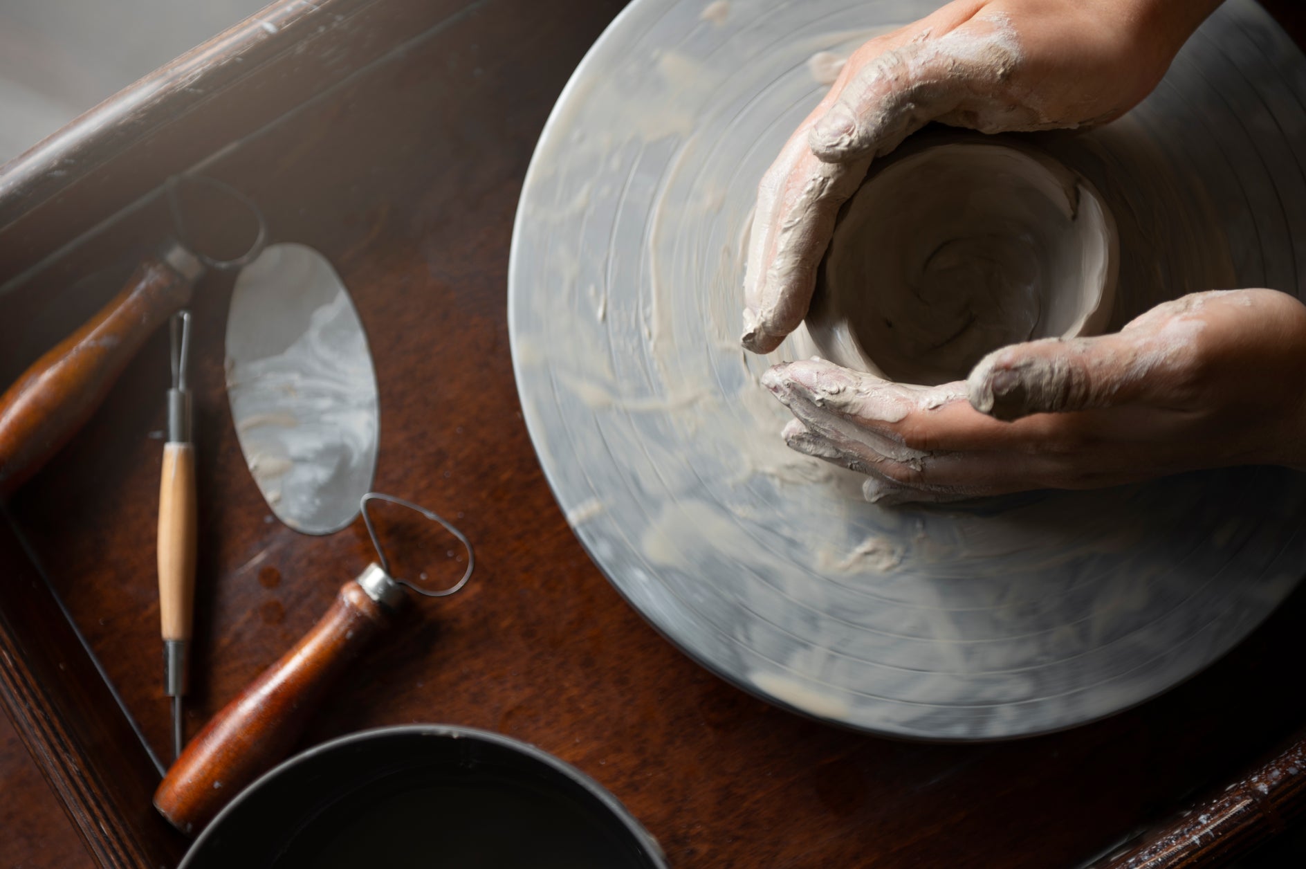 Person working with clay on a pottery wheel, tools on a wooden table.